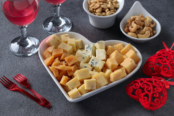 Varieties of elite cheese in a heart-shaped plate, cashews, peanuts and two glasses of wine for Valentine's Day on dark gray background