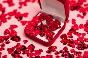 close up of jewelry box with diamond ring near red confetti hearts on pink.