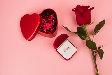 top view of red rose near box with heart-shaped confetti and engagement ring on pink.