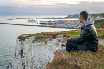 Fototapeta na wymiar Young man exploring the White Cliffs of Dover in UK. Sitting on top of the cliff watching English canal.