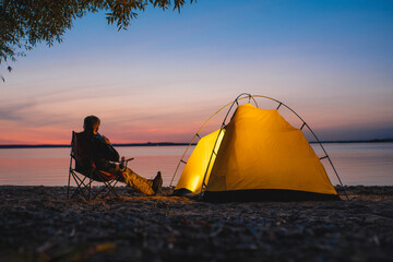 The traveler sits on a camping chair near the glowing tent 