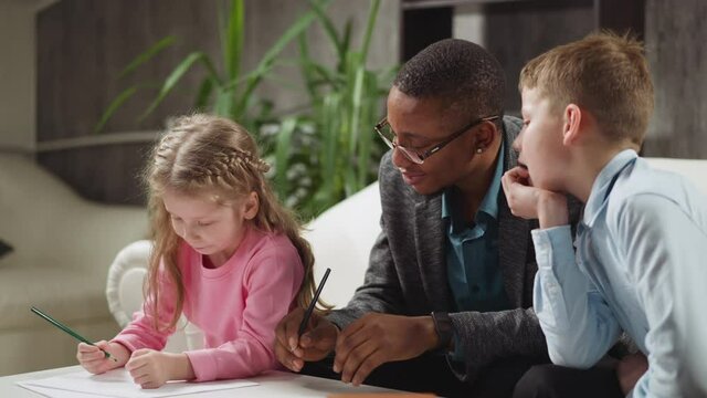 African-American tutor in glasses and schoolboy look peeking at little sister writing letters at home English lesson with native speaker in room slow motion