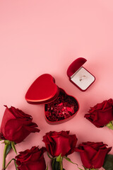 top view of red roses near box with heart-shaped confetti and engagement ring on pink.