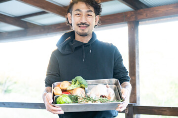 A photo of an Asian (Japanese) man showing vegetables and birds from his farm, looking at the camera.
