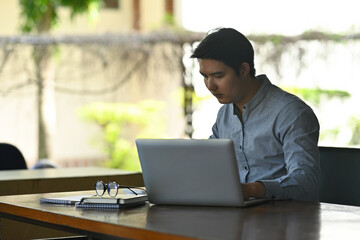 Photo of a young businessman sitting at the wooden working desk and looking at the computer laptop.