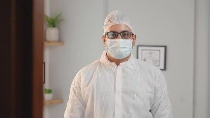 Track in shot of an Indian male or man doctor or a frontline worker protective mask and wearing disposable personal protective equipment kit standing in hospital or clinic and looking at camera