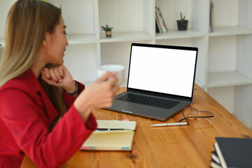 Photo of a white blank screen computer laptop on the wooden table surrounded with blur office woman holds a coffee cup.
