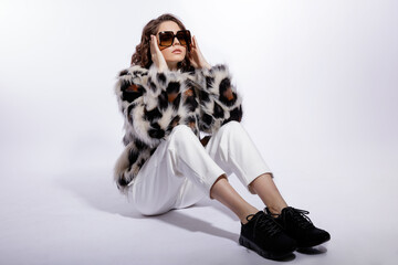 High fashion photo of a beautiful elegant young woman in a pretty leopard print fur coat, stylish sunglasses, trousers, pants, black sneakers posing on white background. Slim figure. Model is sitting