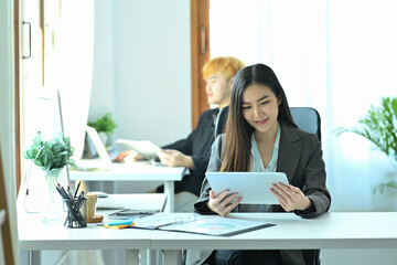 Photo of a young office woman holding a digital tablet at the working desk over the colleague in the office as a background.