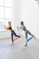 Obraz premium Two smiling athletic women squatting synchronously with outfit in fitness studio. Women are enjoying leg and buttocks work out with resistance band. Training together with friend in gym concept