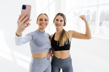 Two sports women meet at gym, making selfie photo while standing together in the white hallway. Meeting friends and having fun at gym