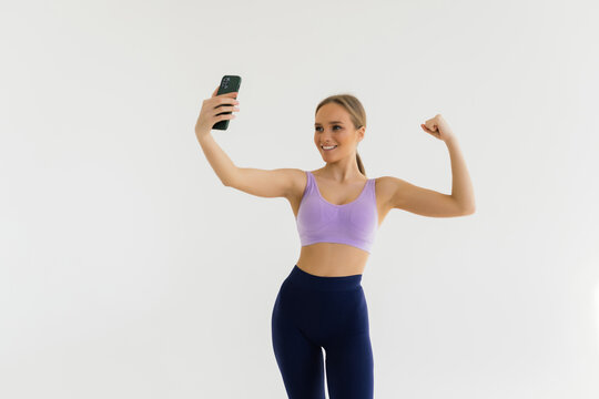 Active and healthy woman in gym taking selfie, showing strong arm, flexing biceps and posing for photo on mobile phone, white background