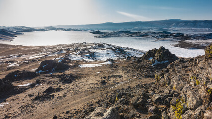 The frozen Siberian lake is surrounded by hills. The snow is lying on the bare ground. A mountain range against a blue sky. In the foreground is a slope of granite rock with yellow lichens. Baikal