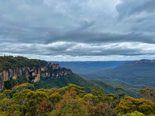 Fototapeta na wymiar The Three Sisters rocks at Blue Mountains with view of dark clouds covering the rainforest trees, Sydney Australia.