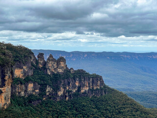 Fototapeta premium The Three Sisters rocks at Blue Mountains with view of dark clouds covering the rainforest trees, Sydney Australia.
