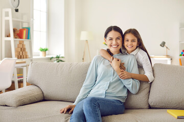 Naklejka na ściany i meble Portrait of smiling young Caucasian mother and small teen daughter relax on couch at home on family weekend. Happy little girl child hug embrace mom show love and care, feel grateful thankful.