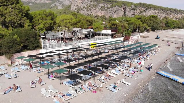 Aerial view of rows of sun loungers with sunbathing tourists in a recreation area on a mountain coast and people swimming in the sea