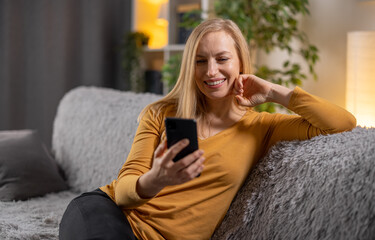 Obraz premium Happy mature woman in casual wear sitting on comfy grey couch and using smartphone. Pretty caucasian lady with blond hair browsing internet on modern gadget.