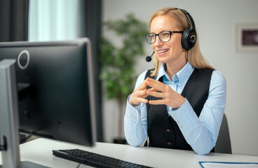 Mature woman sitting at desk in headset and talking during video conference on modern computer....