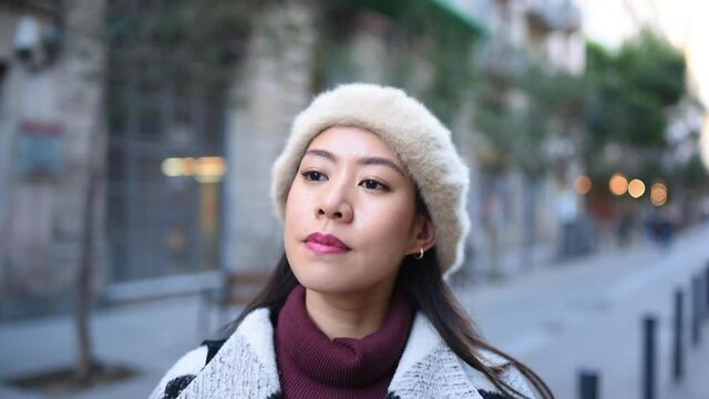 Friendly Asian woman in trendy outerwear and beret taking off face mask and looking at camera with cheerful smile on city street in evening