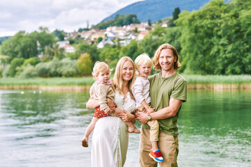 Outdoor portrait of beautiful family, young couple with preschooler boy and toddler girl posing next to lake or river