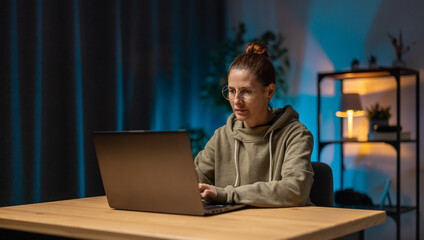 Focused mature woman in eyewear sitting at home office during evening time and working on modern laptop. Freelance, people and technology concept.