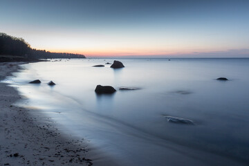 Fototapeta premium Scenic view to the coastal sandy beach with erratic rocks in the water and the sunset colored sea and sky background