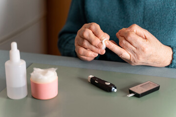 An elderly man wipes the blood on his finger with medical cotton after measuring his blood sugar. Using a glucometer. On the table hydrogen peroxide, lancet device and glucometer.