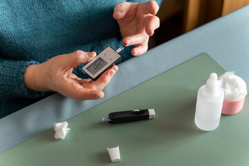 An elderly woman measures her blood sugar level at home with a blood glucose meter. Cotton wool, lancet device and hydrogen peroxide are on the table.