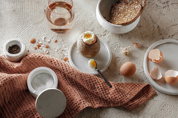 Obraz na płótnie Canvas Breakfast setting with boiled egg in stoneware egg cup, whole grain rye bread in concrete tray, salt flakes and pepper in concrete bowls on rough textured clay background. Healthy breakfast concept
