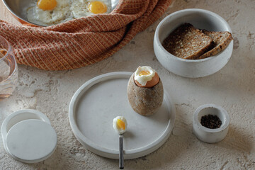 Fried two eggs in copper pan, hard boiled egg in stoneware egg cup, salt flakes, pepper, whole grain rye bread in concrete bowl on rough textured clay background. Healthy breakfast concept.