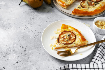 A slice of puff pastry pizza with pear, almonds, gorgonzola ricotta or blue cheese in a white plate on the kitchen table. Delicious layered pie with pear, dorblu, nuts on a light culinary background