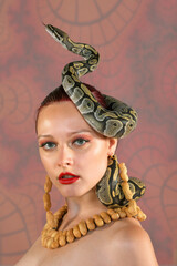A beautiful woman poses nude for the camera. She is 
seen wearing a necklace and earrings made of peanuts. 
A python snake curls itself around her head. The model 
is seen nude in the photograph.
