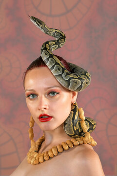 A beautiful woman poses nude for the camera. She is 
seen wearing a necklace and earrings made of peanuts. 
A python snake curls itself around her head. The model 
is seen nude in the photograph.
