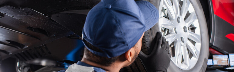 African american mechanic in cap looking at car wheel in garage, banner.