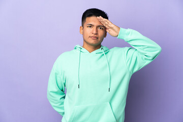 Young Ecuadorian man isolated on purple background saluting with hand with happy expression