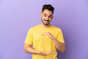 Young caucasian man isolated on purple background holding copyspace imaginary on the palm to insert...