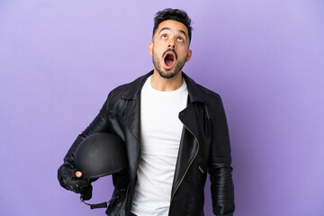 Young man with a motorcycle helmet isolated on purple background looking up and with surprised expression