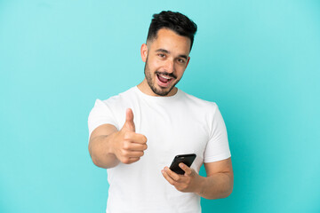 Young caucasian man isolated on blue background using mobile phone while doing thumbs up