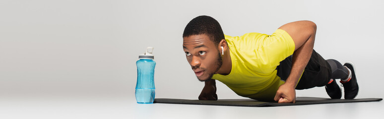 sportive african american man in earphones doing push ups on fitness mat near sports bottle on grey, banner. - obrazy, fototapety, plakaty