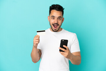 Young caucasian man isolated on blue background buying with the mobile and holding a credit card with surprised expression