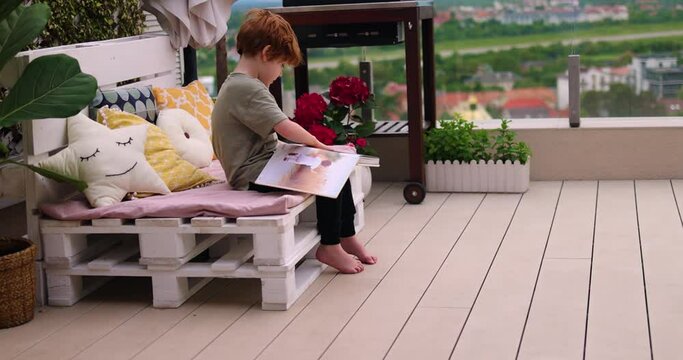 a young boy, cheerful kid flipping the family photo album while sitting on rooftop patio lounge at summer evening