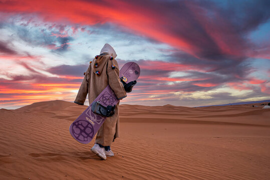 Sahara, Morocco. October 10, 2021. Man in traditional clothes carrying sandboard while walking on sand against cloudy sky during dusk, Man with sandboard walking in desert