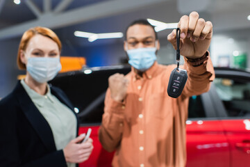 selective focus of key in hand of blurred african american man in medical mask showing success gesture near car dealer.