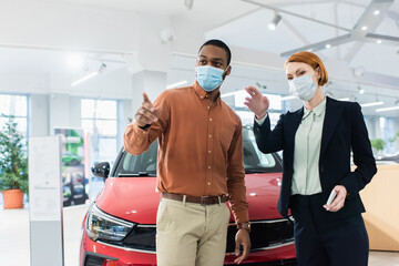 car dealer and african american client in medical masks pointing with fingers in car showroom.