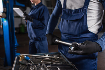 Cropped view of mechanic holding wrench near blurred colleague with digital tablet in garage.