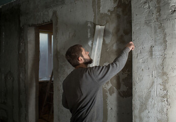Fototapeta na wymiar a man plasterer stitches with a rule at a concrete wall in a room, evaluates its evenness