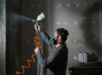 a male plasterer in a construction respirator paints a wall from a spray gun before plastering and...