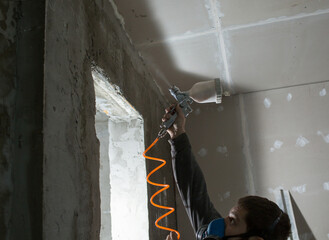 a male plasterer in a construction respirator paints a wall from a spray gun before plastering and putty..