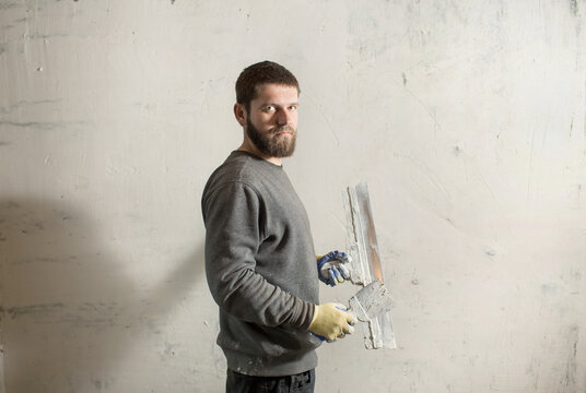 a plasterer man with a beard stands at a rough plastered wall with spatulas